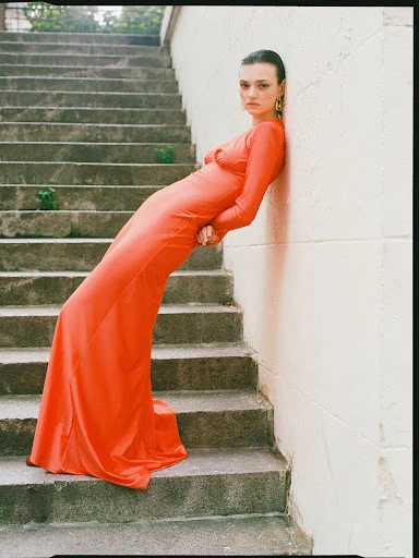 A woman in a long red dress leans against a wall next to a staircase. She wears gold earrings and has her hair slicked back.