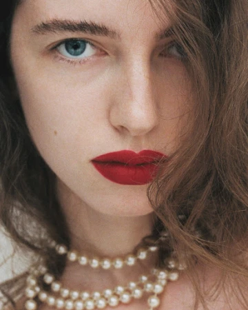Close-up portrait of a woman with red lipstick, blue eyes, and layered pearl necklaces by make-up artist Nolwenn Quintin.