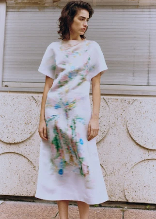 A brunette woman in a painterly white dress stands outdoors against a textured wall. The dress features splashes of blue, green, and pink.