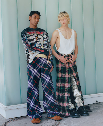 Two young adults pose outdoors in fashionable plaid trousers and unique tops, showcasing style and modern menswear trends.