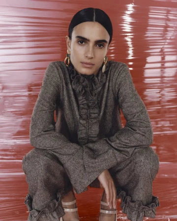 Woman wearing a textured grey ruffled top and trousers posing against a glossy red background.