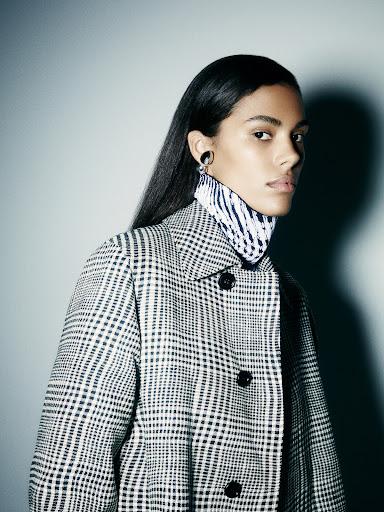 Adult woman with short brown hair wearing makeup and black jacket standing in a photo studio with a grey backdrop.