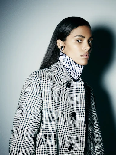 Adult woman with short brown hair wearing makeup and black jacket standing in a photo studio with a grey backdrop.