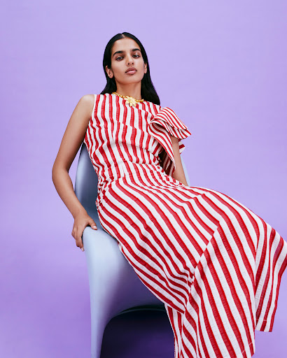 A woman wearing a red and white striped dress poses on a light blue chair in a studio setting with a light purple background.