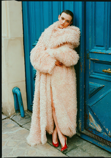 A female model in a pink coat stands against a blue door, wearing red shoes, with an urban environment and a natural light setting.