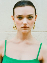 Close-up portrait of a woman with slicked-back dark hair wearing gold drop earrings and a green top against a light wall.