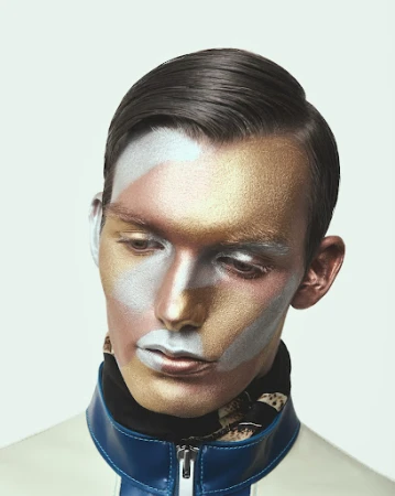 Close-up portrait of a man with metallic gold and silver face makeup wearing a white collared jacket with blue trim.