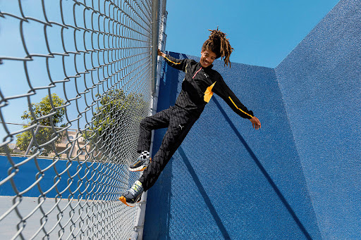 Young man in a black and yellow tracksuit climbs a chain-link fence with blue wall, tree, and sky visible in the background on a sunny day.