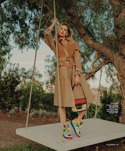 An adult woman with blonde hair poses on a swing wearing a patterned coat with colorful sneakers, a handbag, trees in the background.