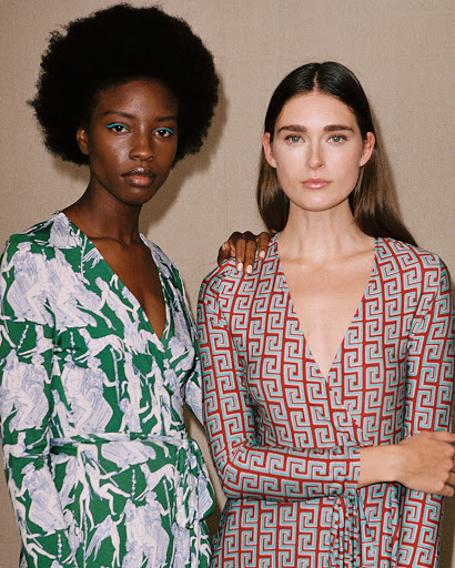 Two women in printed wrap dresses pose in a medium shot. One is wearing a green dress, the other a red dress. Studio lighting.
