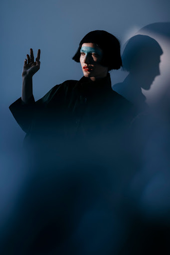 Portrait of an adult female with blue makeup gesturing with her hand, a dark jacket, and bob haircut, captured in a photo studio setting.