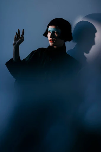Portrait of an adult female with blue makeup gesturing with her hand, a dark jacket, and bob haircut, captured in a photo studio setting.