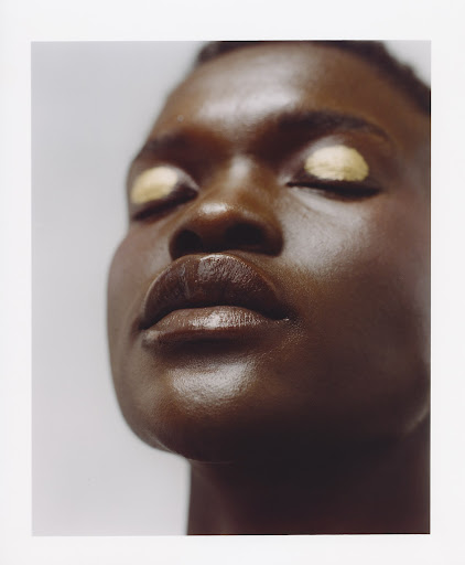 Close-up portrait of a Black woman with dark skin and golden eyeshadow. She has her eyes closed, and the lighting is soft and diffused.