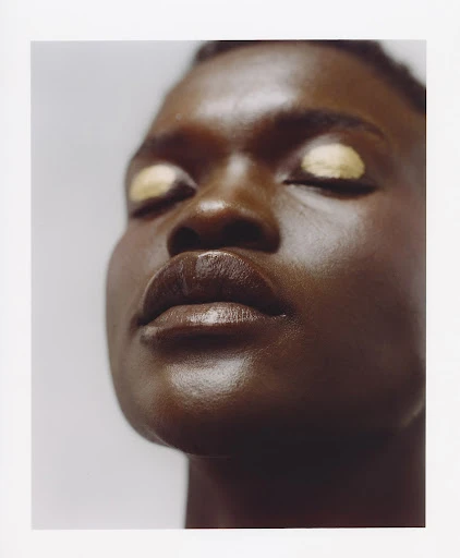 Close-up portrait of a Black woman with dark skin and golden eyeshadow. She has her eyes closed, and the lighting is soft and diffused.
