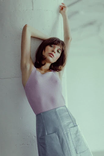 Attractive woman with brown hair leans against a wall wearing a lilac top and metallic blue skirt in an indoor setting with soft lighting.