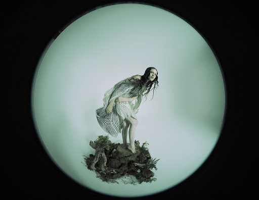 Woman in patterned dress poses on a mossy mound under studio lights in a fisheye lens shot, with a black border.