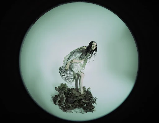 Woman in patterned dress poses on a mossy mound under studio lights in a fisheye lens shot, with a black border.