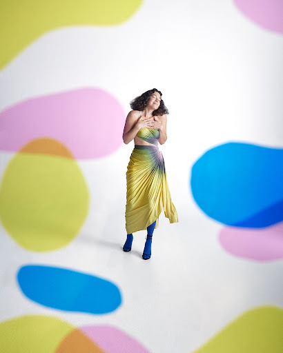 Full-shot of a smiling adult woman in a fashionable yellow and blue skirt and top, posing in a photo studio with colorful bokeh shapes.