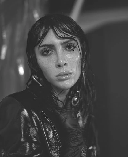 Black and white portrait of a young adult woman with wet hair and a glossy jacket staring intensely into the camera in a studio.