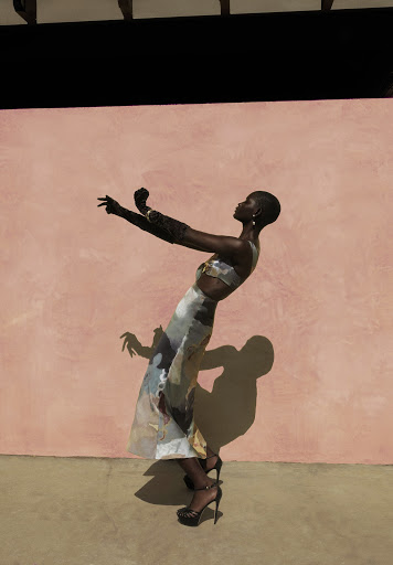 Adult female fashion model poses in a flowing patterned skirt and matching crop top in front of a textured pink wall on tan concrete.
