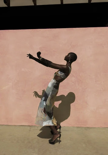 Adult female fashion model poses in a flowing patterned skirt and matching crop top in front of a textured pink wall on tan concrete.