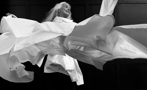 Monochrome portrait of an elderly woman with long white hair wearing a white shirt, arms outstretched in a dance-like pose, against a dark backdrop.