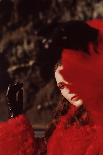 Adult female in red fur coat and black gloves poses outside with dark, textured background and sunlight filtering in from the side.
