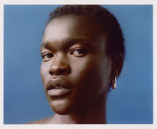 Close-up portrait of a Black person with short dark hair wearing a silver hoop earring against a solid blue background.