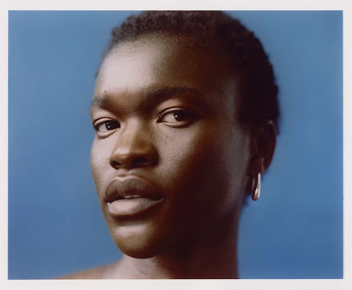 Close-up portrait of a Black person with short dark hair wearing a silver hoop earring against a solid blue background.
