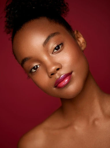 Close-up portrait of a young African American woman with dark hair in a high bun, wearing glossy lip color, against a dark red background.