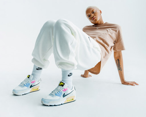Blond woman in a tan t-shirt and white sweatpants poses in front of a white background wearing colorful Nike Air Max 90 sneakers.