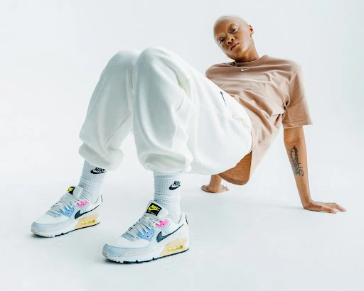 Blond woman in a tan t-shirt and white sweatpants poses in front of a white background wearing colorful Nike Air Max 90 sneakers.