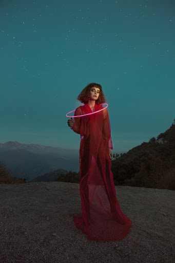 A solitary woman in a red dress stands in a landscape at night, holding a glowing hoop, under a sky full of stars with trees and hills.