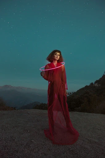 A solitary woman in a red dress stands in a landscape at night, holding a glowing hoop, under a sky full of stars with trees and hills.