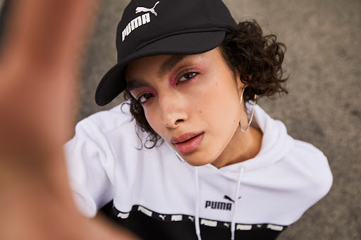 Close-up portrait of a woman wearing a black Puma cap and white hoodie, styled by make-up artist Omayma Ramzy.