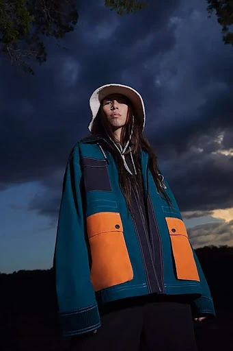 A person wearing a bucket hat and a teal and orange color-block jacket poses in an outdoor setting against a cloudy sky.