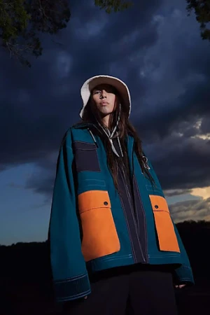 A person wearing a bucket hat and a teal and orange color-block jacket poses in an outdoor setting against a cloudy sky.