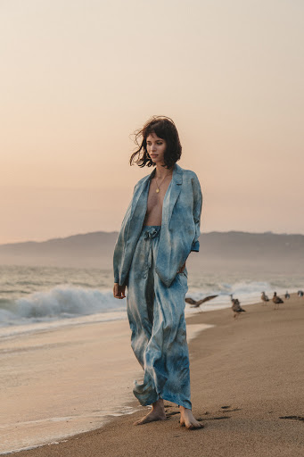 A woman in a blue patterned pantsuit standing barefoot on a beach at sunset near the ocean, with seagulls in the background.