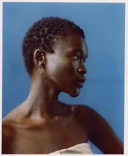 Portrait of a dark-skinned woman with short hair, wearing a white top against a solid blue backdrop, and gold earrings.