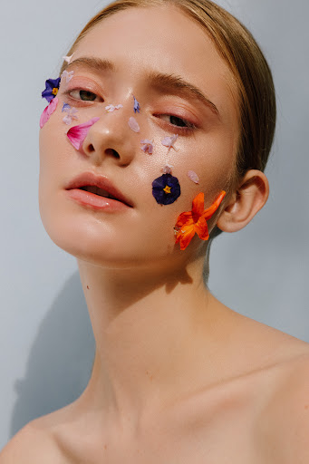 Close-up portrait of a fair-skinned woman with colorful flower petals artfully placed on her face, against a light blue background.