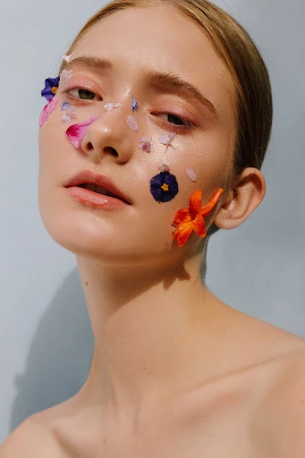 Close-up portrait of a fair-skinned woman with colorful flower petals artfully placed on her face, against a light blue background.
