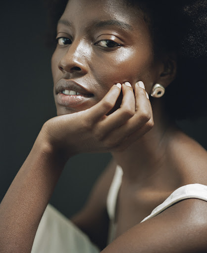 Close-up portrait of a Black woman with natural hair, hand on face, wearing stud earrings against a dark background, beauty shot.