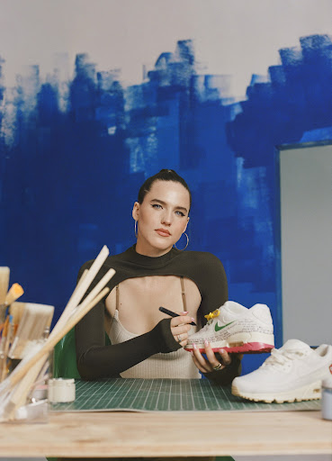 Woman painting a Nike Air Max 90 sneaker with a paintbrush in an indoor studio setting with a blue abstract background painting.