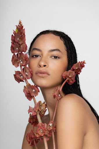 Portrait of a woman with braided hair partially covered by red flowers. Light skin, neutral expression, studio lighting.