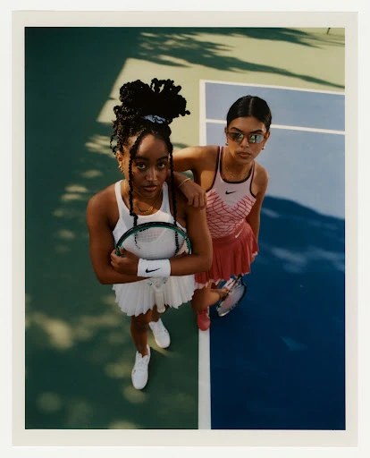 Two young women pose on a tennis court, one resting a hand on the other's shoulder. Both wear tennis outfits and carry tennis rackets.