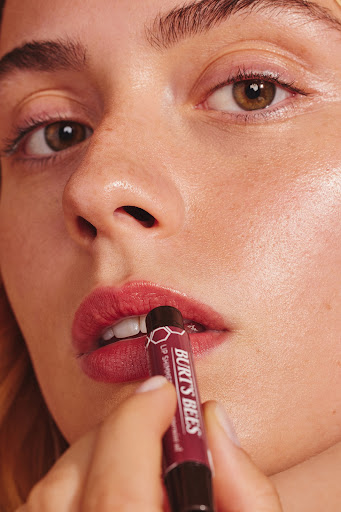 Close-up shot of an adult female applying deep red lipstick from Burt's Bees. The camera focuses on the model's face and hands.