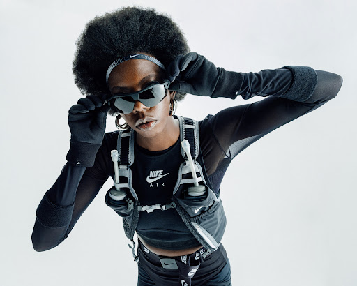 Athletic woman in Nike sports apparel and sunglasses adjusts eyewear against a bright background, showcasing fitness and style.