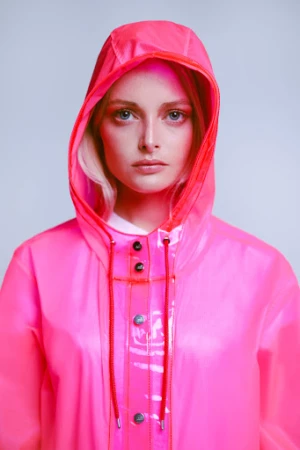 Portrait of a woman wearing a bright pink hooded raincoat with dark buttons against a neutral background.