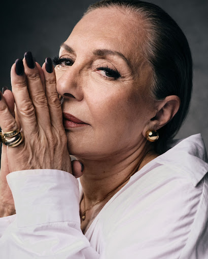 Portrait of an adult woman with fair skin and short dark hair wearing a white collared shirt and gold jewelry against gray background.