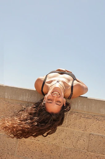 Upside-down view of a smiling woman with long brown hair hanging over a wall under a clear blue sky in an outdoor setting.